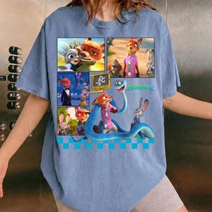 May include: Light blue t-shirt featuring a colorful graphic from the animated movie "Zootopia 2". The graphic is a collage of images with the characters Nick Wilde, Judy Hopps, and others. The text "Zootopia 2" is also visible.