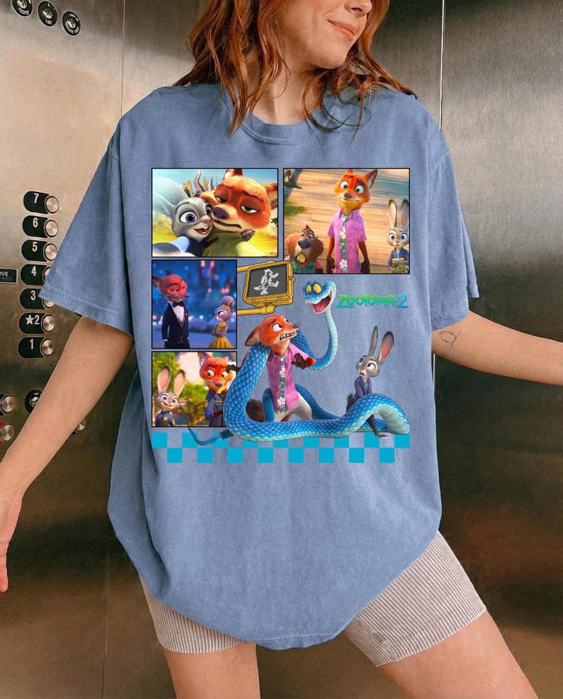 May include: Light blue t-shirt featuring a colorful graphic from the animated movie "Zootopia 2". The graphic is a collage of images with the characters Nick Wilde, Judy Hopps, and others. The text "Zootopia 2" is also visible.