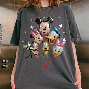 May include: Dark gray t-shirt featuring a colorful graphic of Mickey Mouse, Minnie Mouse, Donald Duck, Daisy Duck, Goofy, and Pluto as balloon characters. The design includes star accents and red string details.