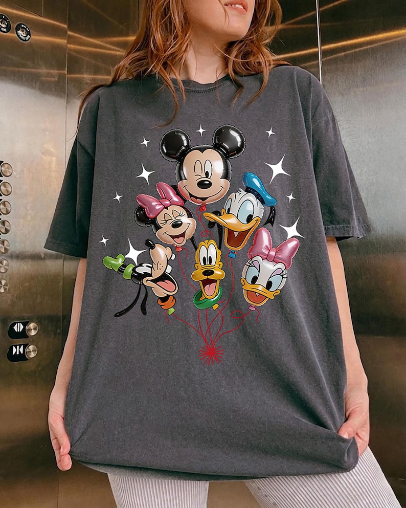 May include: Dark gray t-shirt featuring a colorful graphic of Mickey Mouse, Minnie Mouse, Donald Duck, Daisy Duck, Goofy, and Pluto as balloon characters. The design includes star accents and red string details.