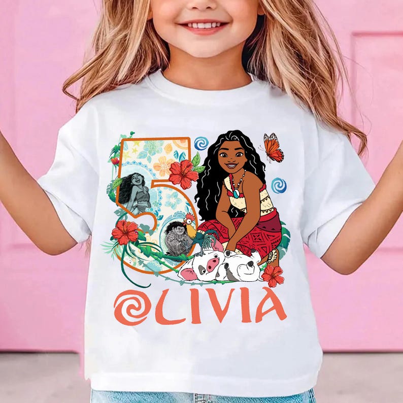 May include: White t-shirt featuring a colorful graphic of a cartoon character, flowers, and the number 5. The name "OLIVIA" is printed in orange script below the graphic. The shirt is designed for a child.