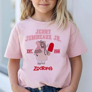 May include: A light pink t-shirt featuring the text "JERRY JUMBEAUX JR." above an illustration of an elephant holding a red popsicle. The shirt also includes the text "EST. 2016" and "ZOOTOPIA" in a red font.