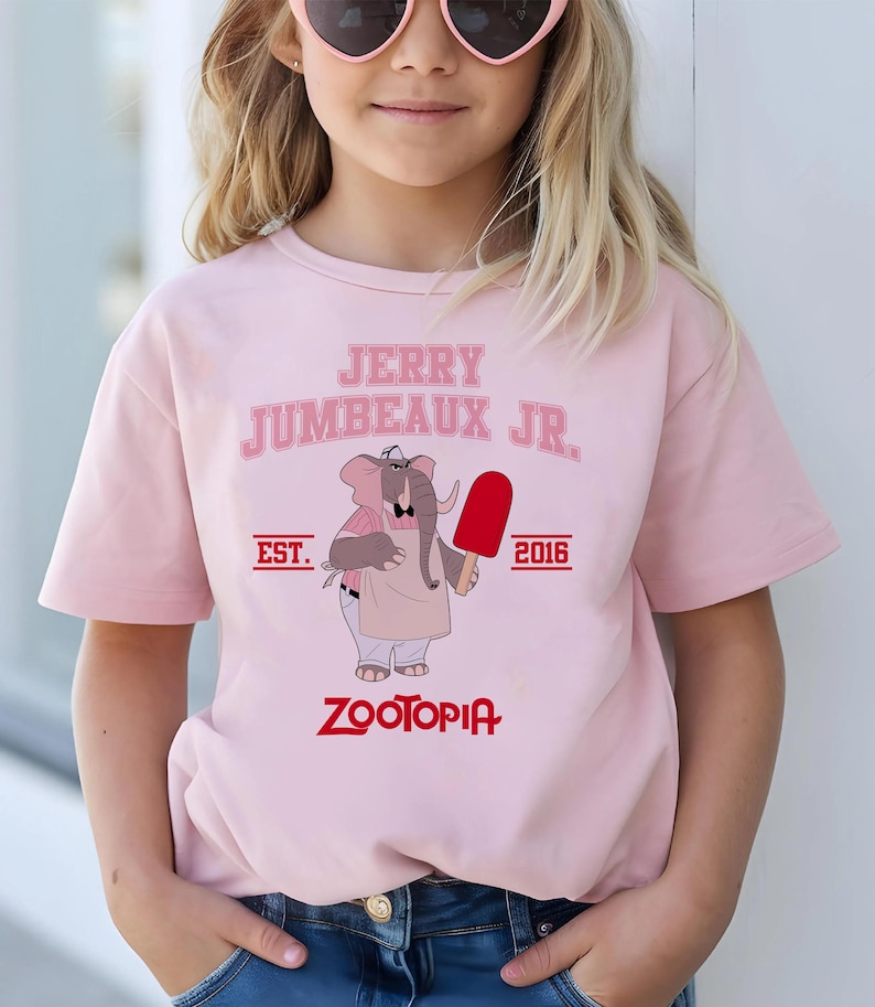 May include: A light pink t-shirt featuring the text "JERRY JUMBEAUX JR." above an illustration of an elephant holding a red popsicle. The shirt also includes the text "EST. 2016" and "ZOOTOPIA" in a red font.