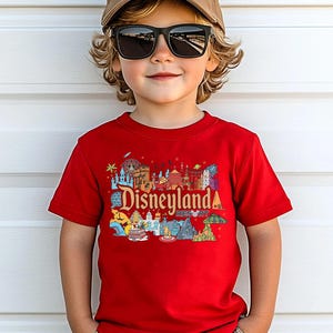 Op de afbeelding: Een kind draagt een rood t-shirt met een kleurrijke Disneyland Resort-afbeelding. Het shirt toont verschillende iconische bezienswaardigheden en het Disneyland-logo. Het kind draagt ook een bruine pet en een zwarte zonnebril.