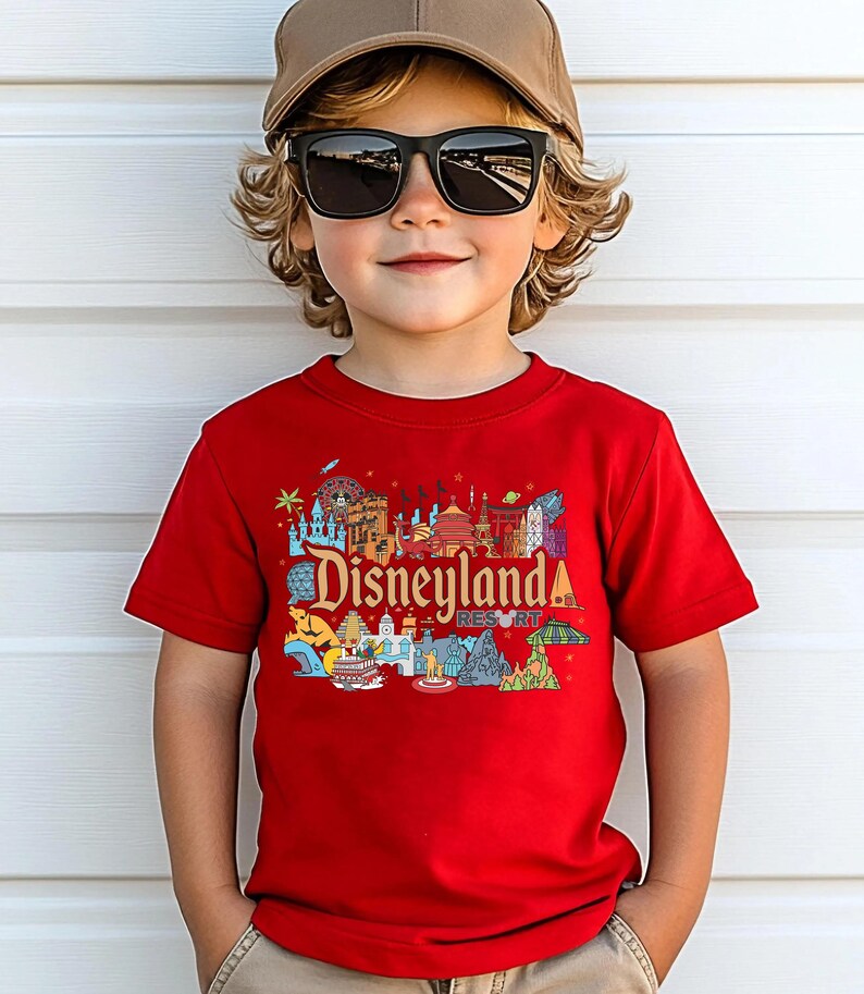 Op de afbeelding: Een kind draagt een rood t-shirt met een kleurrijke Disneyland Resort-afbeelding. Het shirt toont verschillende iconische bezienswaardigheden en het Disneyland-logo. Het kind draagt ook een bruine pet en een zwarte zonnebril.