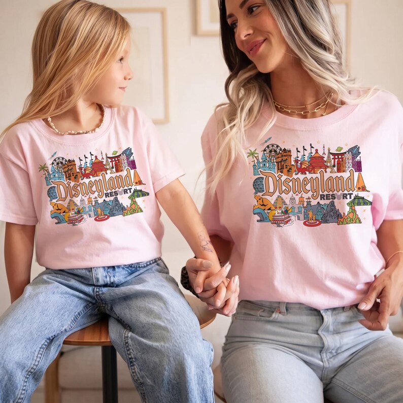 Op de afbeelding: Twee roze T-shirts met een Disneyland Resort-afbeelding. De afbeelding bevat kleurrijke illustraties van verschillende bezienswaardigheden en personages. De shirts worden gedragen door een moeder en kind, beiden dragen een blauwe jeans.
