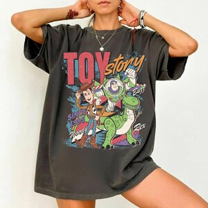 May include: Dark gray t-shirt featuring the words "Toy Story" in red and orange, with Woody, Buzz Lightyear, and Rex the dinosaur. The shirt also includes the text "Since 1995".