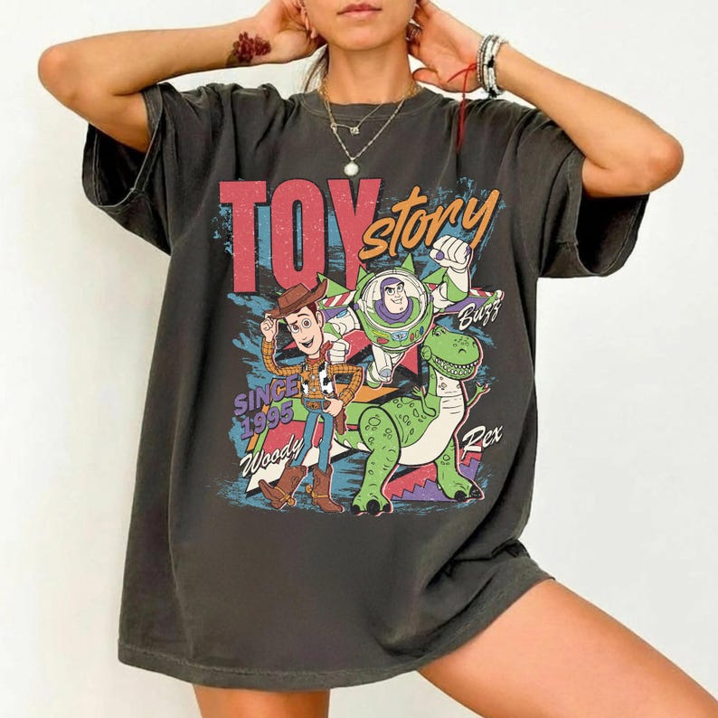 May include: Dark gray t-shirt featuring the words "Toy Story" in red and orange, with Woody, Buzz Lightyear, and Rex the dinosaur. The shirt also includes the text "Since 1995".