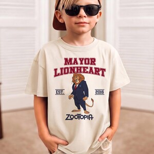 May include: A beige t-shirt featuring the text "MAYOR LIONHEART" in red, with an illustration of a lion in a suit. Below the illustration is the word "ZOOTOPIA". The shirt also has "EST. 2016" printed on it.