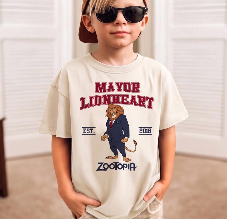 May include: A beige t-shirt featuring the text "MAYOR LIONHEART" in red, with an illustration of a lion in a suit. Below the illustration is the word "ZOOTOPIA". The shirt also has "EST. 2016" printed on it.