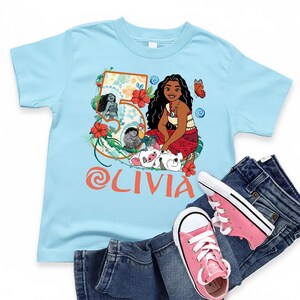 May include: A light blue t-shirt featuring a colorful graphic of a cartoon character, flowers, and the name "Olivia" in orange. The shirt is paired with blue jeans and pink sneakers.