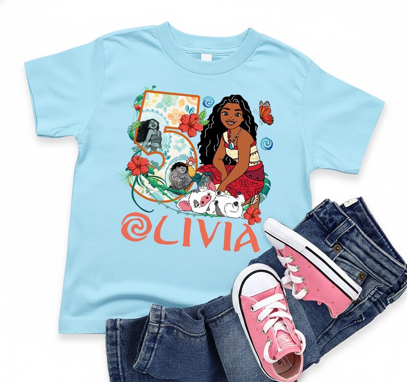May include: A light blue t-shirt featuring a colorful graphic of a cartoon character, flowers, and the name "Olivia" in orange. The shirt is paired with blue jeans and pink sneakers.