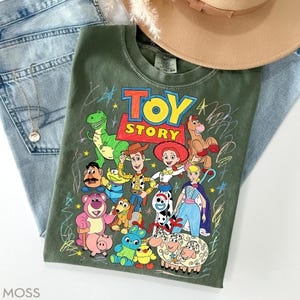 May include: Olive green t-shirt featuring a colorful graphic of characters from the animated film "Toy Story." The design includes Woody, Buzz Lightyear, and other beloved characters. The shirt is paired with blue jeans and a tan hat.