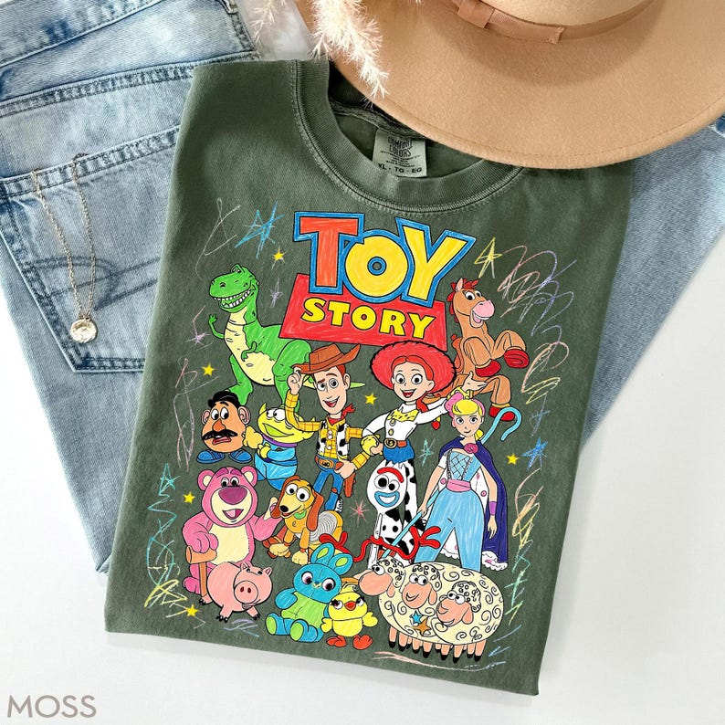 May include: Olive green t-shirt featuring a colorful graphic of characters from the animated film "Toy Story." The design includes Woody, Buzz Lightyear, and other beloved characters. The shirt is paired with blue jeans and a tan hat.