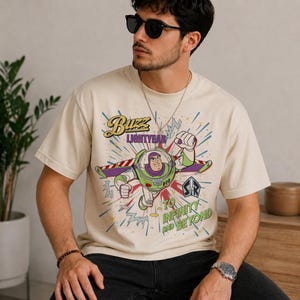 May include: Cream-coloured t-shirt with a graphic of Buzz Lightyear. The design shows Buzz in his spacesuit, with the words "Buzz Lightyear" and "To Infinity and Beyond" in a colourful, retro style. The shirt has a crew neck.