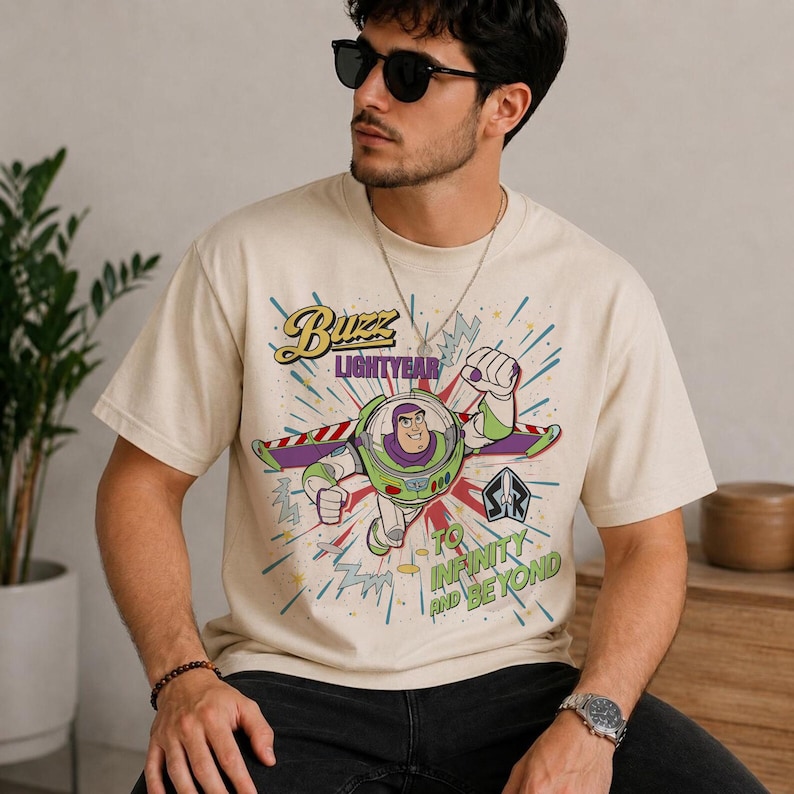 May include: Cream-coloured t-shirt with a graphic of Buzz Lightyear. The design shows Buzz in his spacesuit, with the words "Buzz Lightyear" and "To Infinity and Beyond" in a colourful, retro style. The shirt has a crew neck.