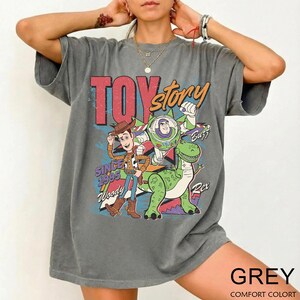 May include: A gray t-shirt featuring a colorful graphic of characters from the movie "Toy Story." The design includes Woody, Buzz Lightyear, and Rex, with the text "TOY STORY" and "SINCE 1995." The shirt is labeled "GREY" and "COMFORT COLORT."