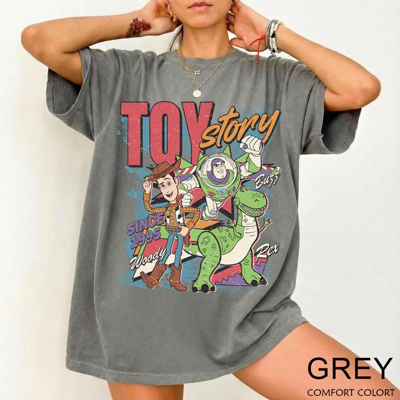 May include: A gray t-shirt featuring a colorful graphic of characters from the movie "Toy Story." The design includes Woody, Buzz Lightyear, and Rex, with the text "TOY STORY" and "SINCE 1995." The shirt is labeled "GREY" and "COMFORT COLORT."