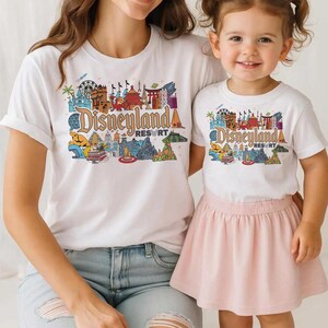 Op de afbeelding: Witte T-shirts met een kleurrijke "Disneyland Resort" afbeelding. De afbeelding toont verschillende bezienswaardigheden en attracties. E&eacute;n shirt wordt gedragen door een volwassene, de andere door een kind, gecombineerd met een roze rok en een blauwe jeans.