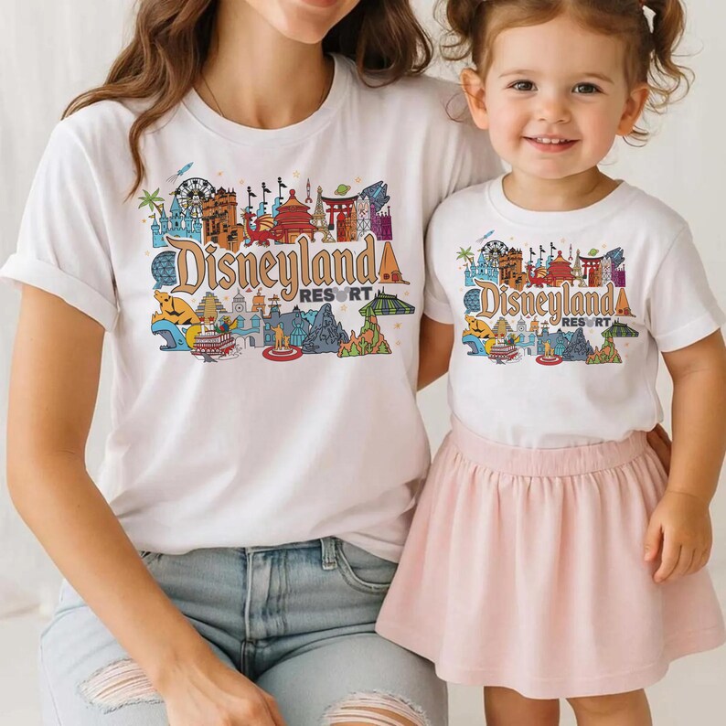 Op de afbeelding: Witte T-shirts met een kleurrijke "Disneyland Resort" afbeelding. De afbeelding toont verschillende bezienswaardigheden en attracties. E&eacute;n shirt wordt gedragen door een volwassene, de andere door een kind, gecombineerd met een roze rok en een blauwe jeans.