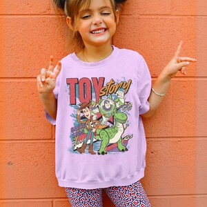 May include: A light purple sweatshirt featuring the words "Toy Story" in red and green, with Woody, Buzz Lightyear, and Rex the dinosaur. The sweatshirt has the text "Since 1995" and "Woody" printed on it.