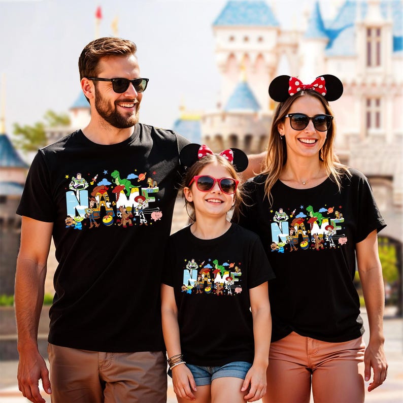 May include: Three people wearing black t-shirts with a colorful graphic design featuring the word "NAME" and cartoon characters. The shirts are paired with sunglasses and accessories. The background includes a castle.