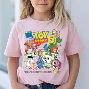 May include: A light pink t-shirt featuring a colorful graphic of characters from the animated film "Toy Story 5". The design includes the text "TOY STORY" and "YOU'VE GOT A FRIEND IN ME". The shirt is worn by a person.