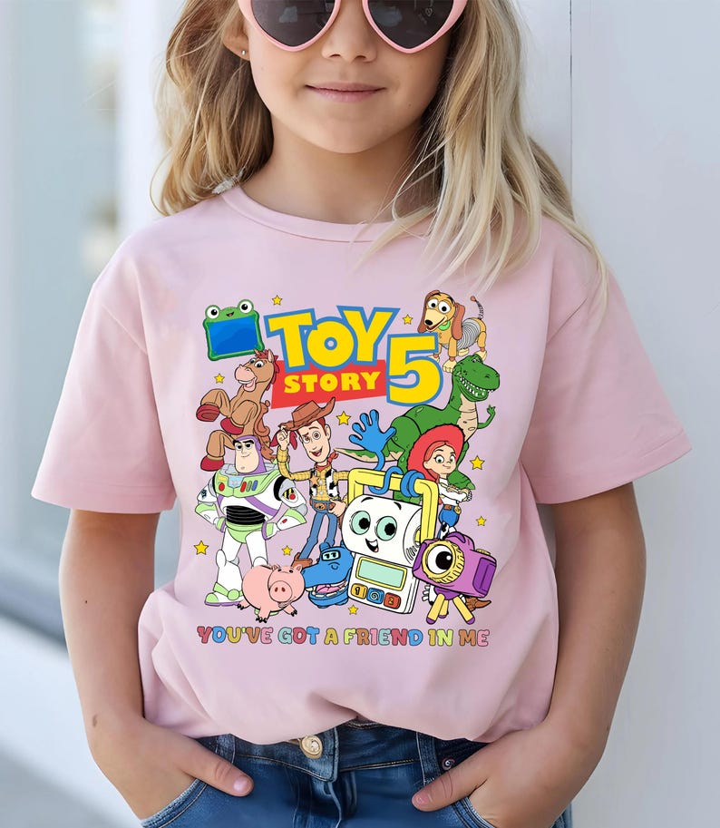 May include: A light pink t-shirt featuring a colorful graphic of characters from the animated film "Toy Story 5". The design includes the text "TOY STORY" and "YOU'VE GOT A FRIEND IN ME". The shirt is worn by a person.