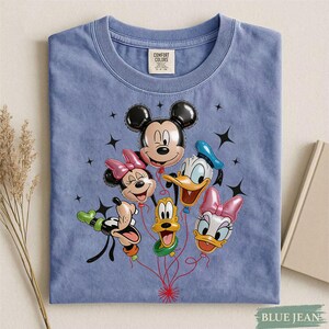 May include: A blue Comfort Colors t-shirt featuring a graphic of Mickey Mouse, Minnie Mouse, Donald Duck, Daisy Duck, Goofy, and Pluto as balloon characters. The design includes black star accents and red balloon strings.