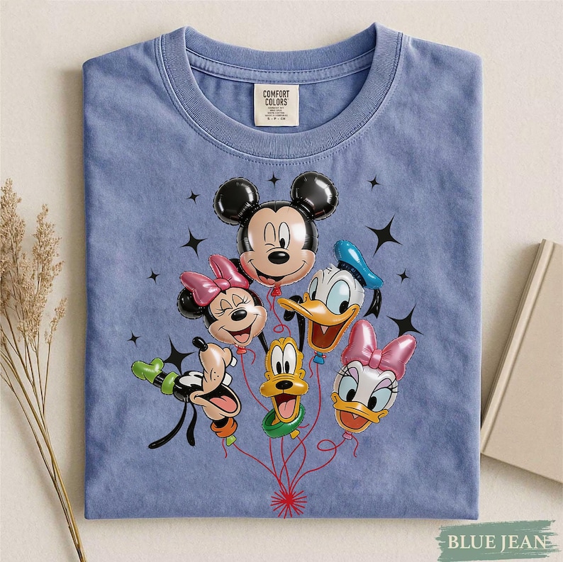 May include: A blue Comfort Colors t-shirt featuring a graphic of Mickey Mouse, Minnie Mouse, Donald Duck, Daisy Duck, Goofy, and Pluto as balloon characters. The design includes black star accents and red balloon strings.