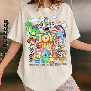 May include: Cream-colored t-shirt featuring a colorful graphic of Toy Story characters, including Woody, Buzz Lightyear, and Rex. The words "TOY STORY" and "YOU'VE GOT A FRIEND IN ME" are also printed on the shirt.