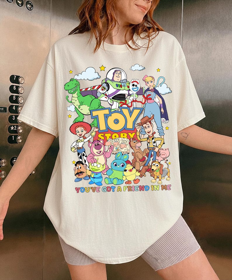 May include: Cream-colored t-shirt featuring a colorful graphic of Toy Story characters, including Woody, Buzz Lightyear, and Rex. The words "TOY STORY" and "YOU'VE GOT A FRIEND IN ME" are also printed on the shirt.