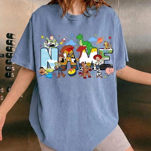 May include: A light blue t-shirt featuring the word "NAME" in a playful, colorful design. The letters are filled with cartoon characters, including a dinosaur, a cowboy, and a space ranger. Stars and planets are scattered around the letters.