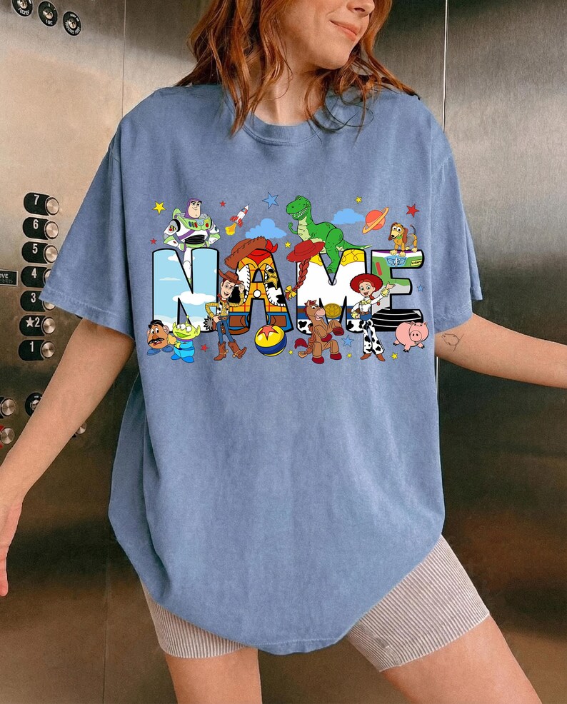 May include: A light blue t-shirt featuring the word "NAME" in a playful, colorful design. The letters are filled with cartoon characters, including a dinosaur, a cowboy, and a space ranger. Stars and planets are scattered around the letters.