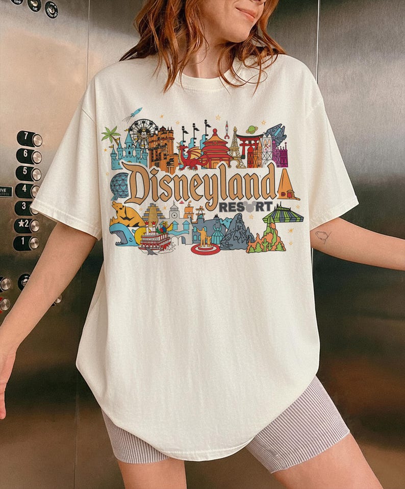 Op de afbeelding: Cr&egrave;mekleurig T-shirt met een kleurrijke Disneyland Resort-afbeelding. Het ontwerp bevat iconische bezienswaardigheden en het Disneyland-logo in vintage stijl. Het shirt heeft korte mouwen en een relaxte pasvorm.