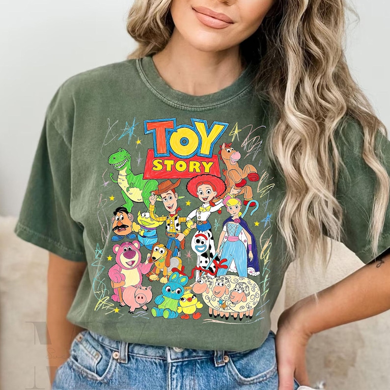 May include: Olive green t-shirt featuring a colorful graphic of characters from the animated film "Toy Story." The design includes Woody, Buzz Lightyear, Jessie, and other characters. The words "Toy Story" are in a bold, colorful font.