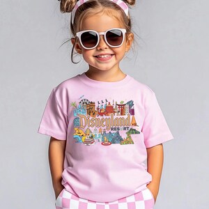 Op de afbeelding: Een kind draagt een lichtroze t-shirt met een kleurrijke Disneyland Resort-afbeelding. Het shirt heeft verschillende Disneyland-bezienswaardigheden. Het kind draagt ook een zonnebril en een roze en witte haarband.