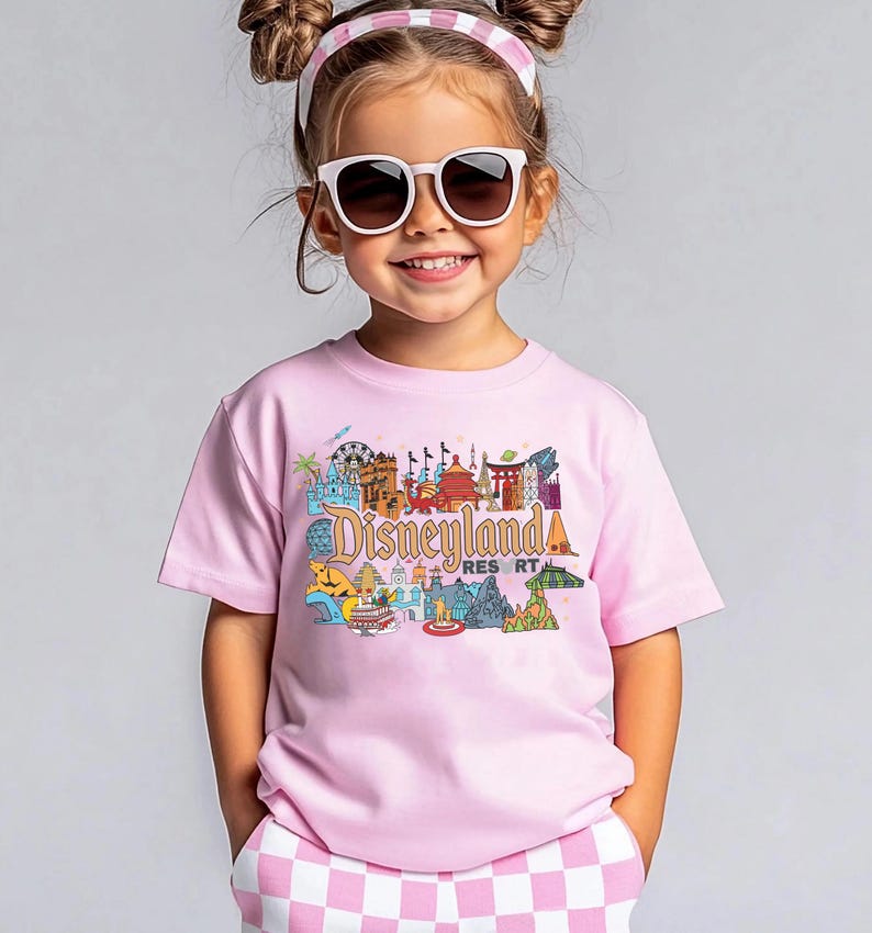 Op de afbeelding: Een kind draagt een lichtroze t-shirt met een kleurrijke Disneyland Resort-afbeelding. Het shirt heeft verschillende Disneyland-bezienswaardigheden. Het kind draagt ook een zonnebril en een roze en witte haarband.