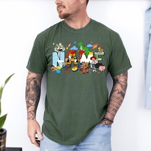 May include: Green t-shirt featuring the word "NAME" in colorful letters with cartoon characters. The design includes characters like Buzz Lightyear, Woody, and Rex. The shirt is a comfortable, casual style.