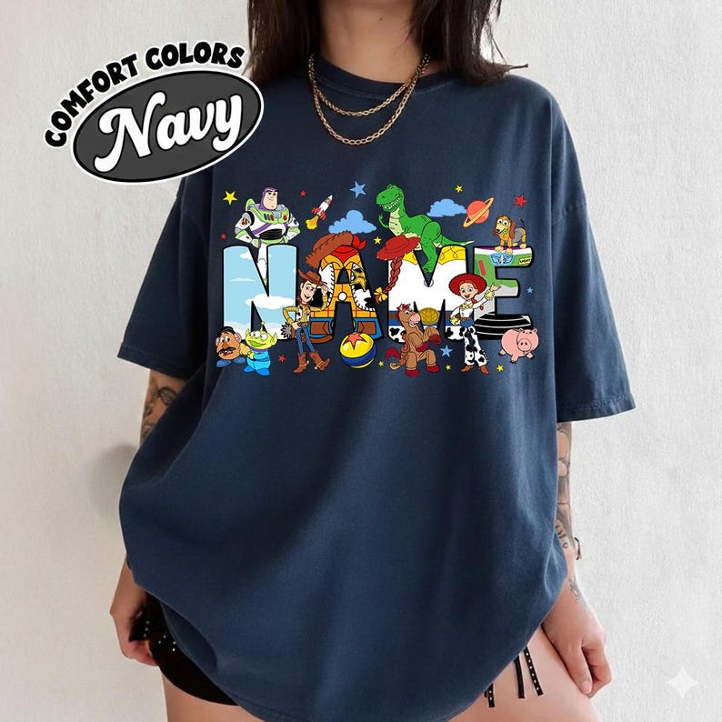 May include: Navy blue t-shirt with the word "NAME" in colorful cartoon character designs. The shirt features the text "COMFORT COLORS" and "Navy" in a white oval. The design includes characters from a popular animated movie.