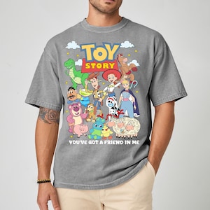 May include: Gray t-shirt featuring a colorful graphic of characters from the animated film "Toy Story." The design includes Woody, Buzz Lightyear, and other beloved characters, with the text "TOY STORY" and "YOU'VE GOT A FRIEND IN ME."
