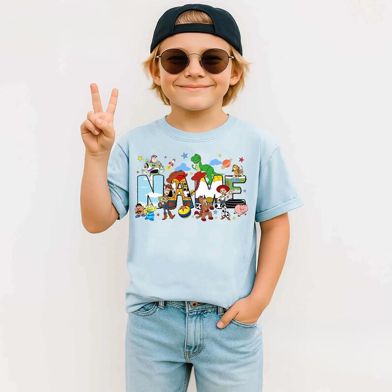 May include: A light blue t-shirt featuring the word "NAME" in colorful block letters, with cartoon characters from a popular animated movie. The shirt is worn by a child wearing sunglasses and a black cap.