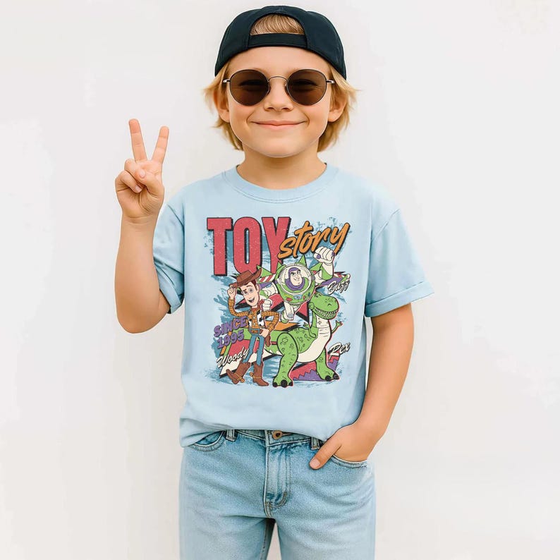 May include: A light blue t-shirt featuring the words "Toy Story" in red and yellow, with Woody, Buzz Lightyear, and Rex the dinosaur. The shirt is paired with light blue jeans and a black cap.