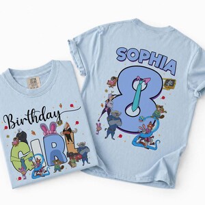 May include: Two light blue t-shirts with cartoon graphics. One shirt says "Birthday Girl" with cartoon characters. The other shirt says "Sophia 8" with cartoon characters. Both shirts are made of cotton.