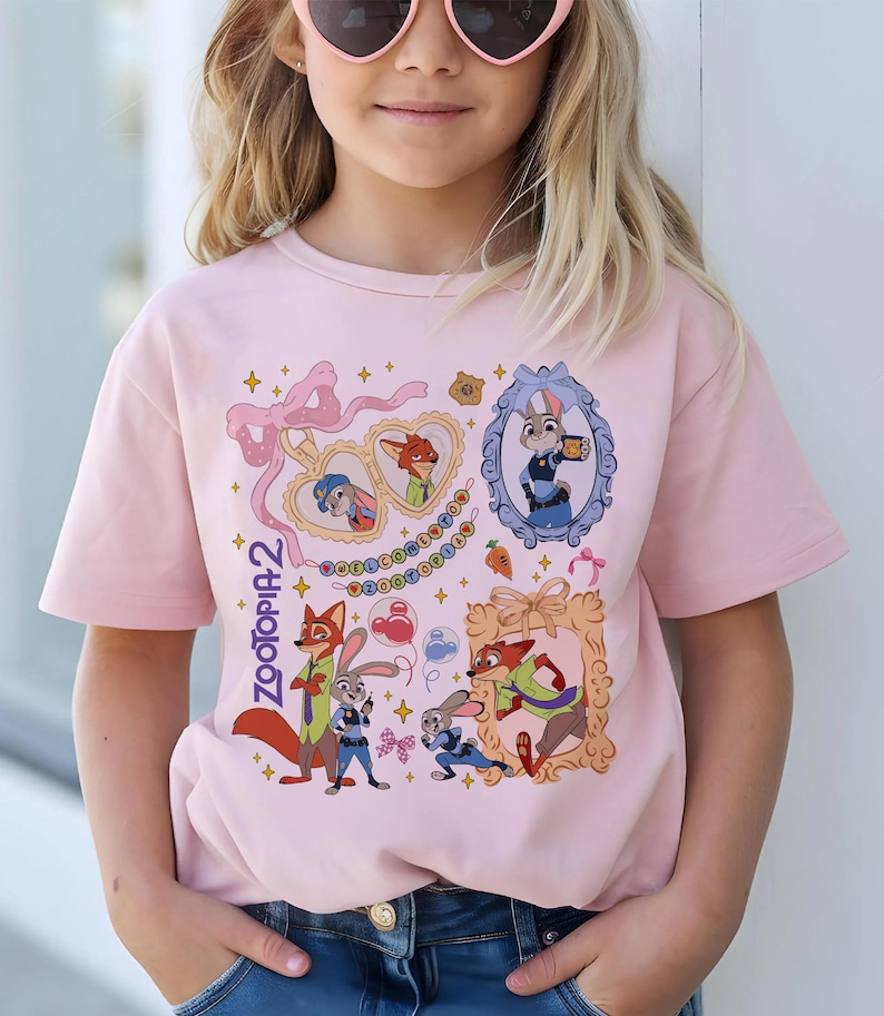 May include: A light pink t-shirt featuring characters from the movie Zootopia 2. The design includes framed portraits of Judy Hopps and Nick Wilde, along with other characters and decorative elements. The text "Zootopia 2" is also visible.