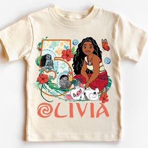 May include: Cream-colored t-shirt featuring a cartoon illustration of a character with long black hair, a red skirt, and a necklace. The number "5" is decorated with flowers and the name "OLIVIA" is printed in orange. A butterfly and other characters are also featured.