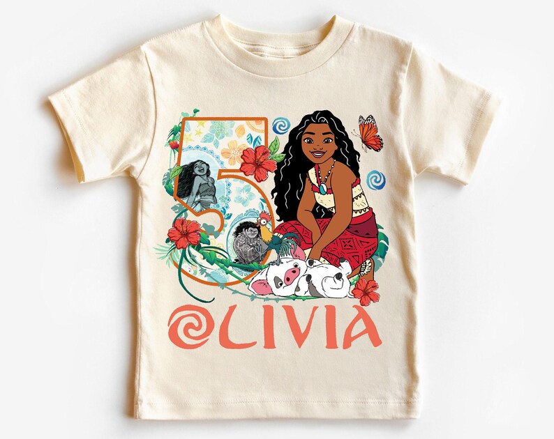 May include: Cream-colored t-shirt featuring a cartoon illustration of a character with long black hair, a red skirt, and a necklace. The number "5" is decorated with flowers and the name "OLIVIA" is printed in orange. A butterfly and other characters are also featured.