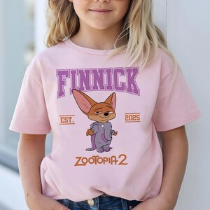 K&ouml;nnte beinhalten: Hellrosa T-Shirt mit dem Charakter Finnick aus Zootopia 2. Das Design zeigt den Namen "FINNICK" in lila Buchstaben, den Text "EST. 2025" und den Filmtitel "ZOOTOPIA 2" in Orange.