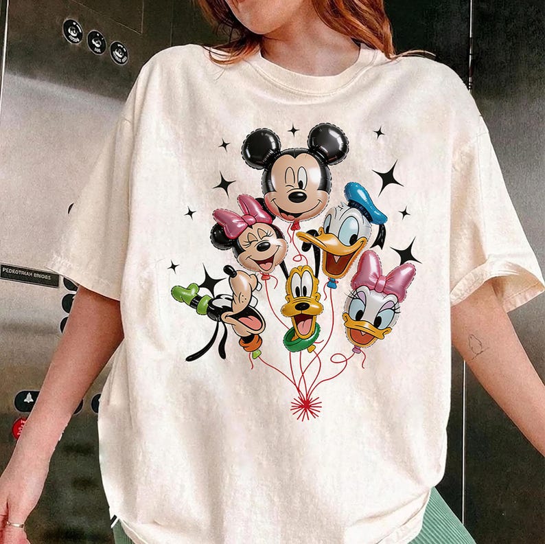 May include: Cream-colored t-shirt featuring a graphic of Mickey Mouse, Minnie Mouse, Donald Duck, Daisy Duck, and Goofy as balloon characters. The balloons are connected by red strings, with black star accents. The shirt is a casual style.