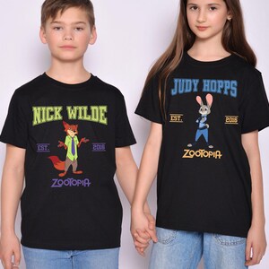 May include: Black t-shirts featuring characters from the animated film Zootopia. One shirt displays "Nick Wilde" with a fox graphic, the other says "Judy Hopps" with a bunny graphic. Both shirts have "Zootopia" and the year "2016".